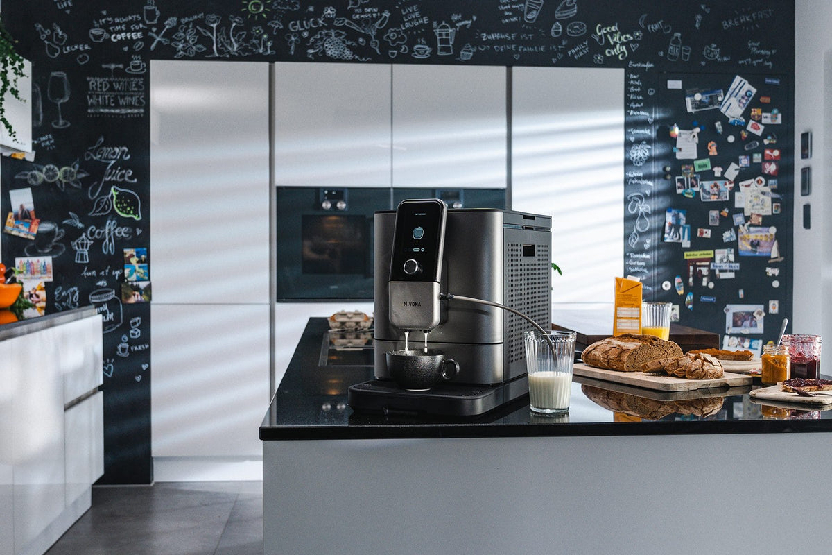 NIVONA NIVO 8103 Kaffeevollautomat Titan auf einer Kücheninsel, daneben ein gedeckter Tisch mit Brot und Saft