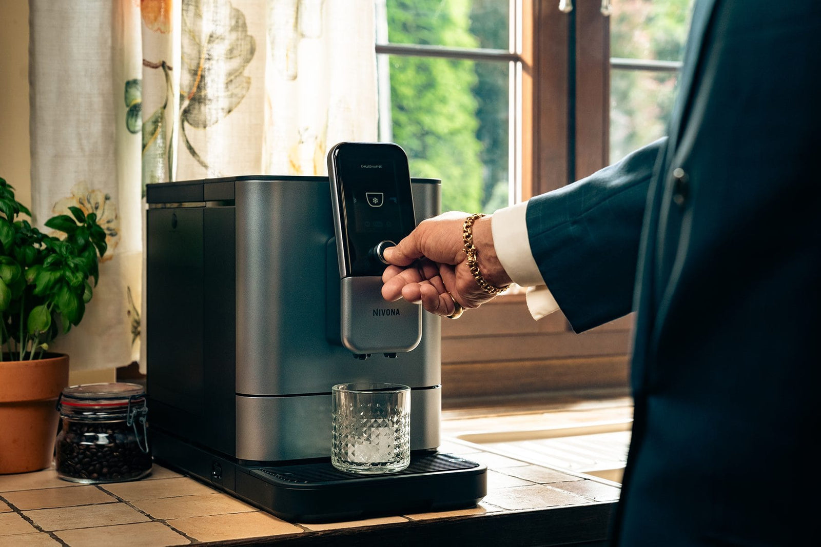 NIVONA NIVO 8'107 am sonnigen Küchenfenster, eine Person nimmt ein Glas mit gekühltem Kaffee entgegen