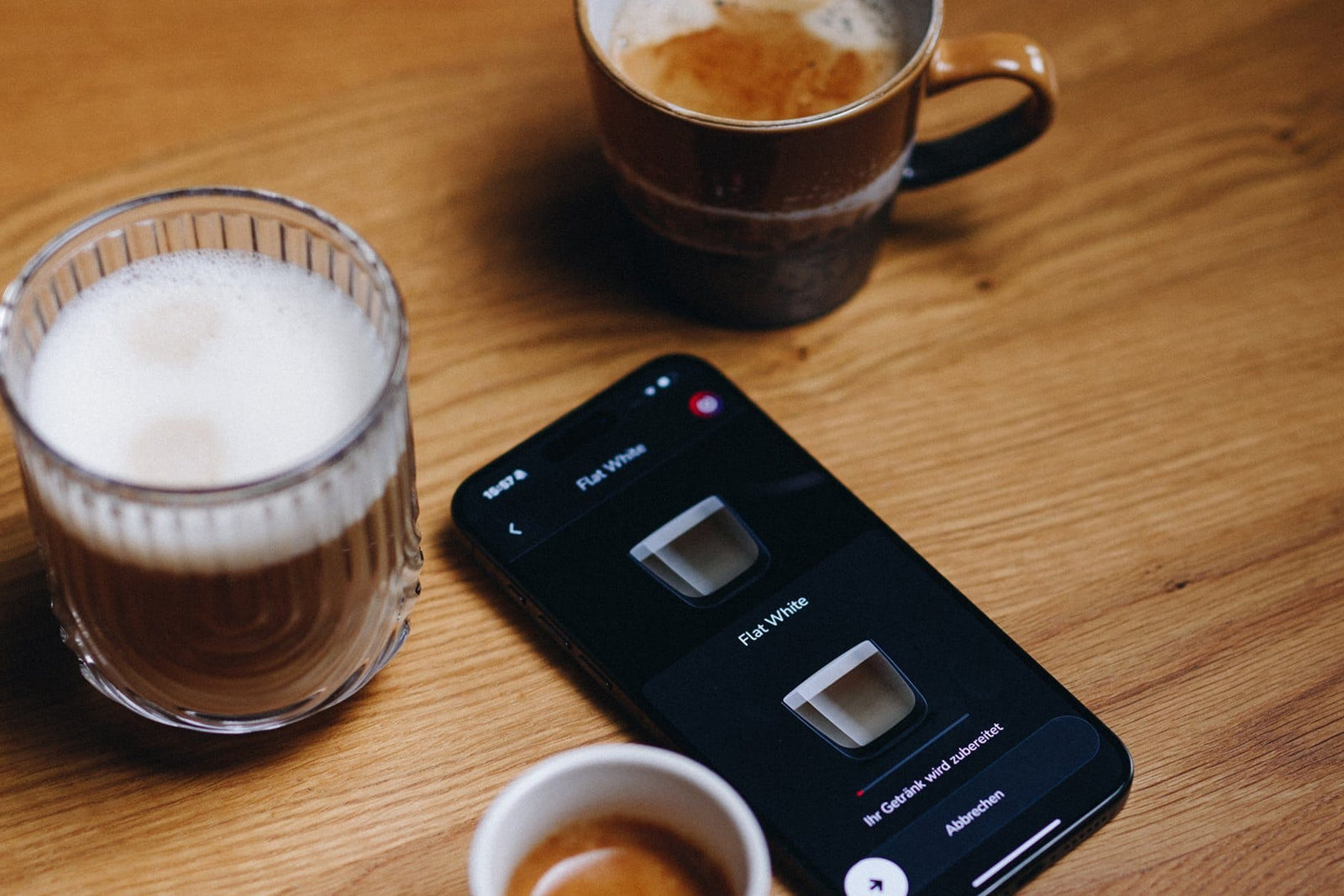 NIVONA App auf dem Smartphone startet einen Flat White, daneben stehen Latte Macchiato, Kaffee und Espresso