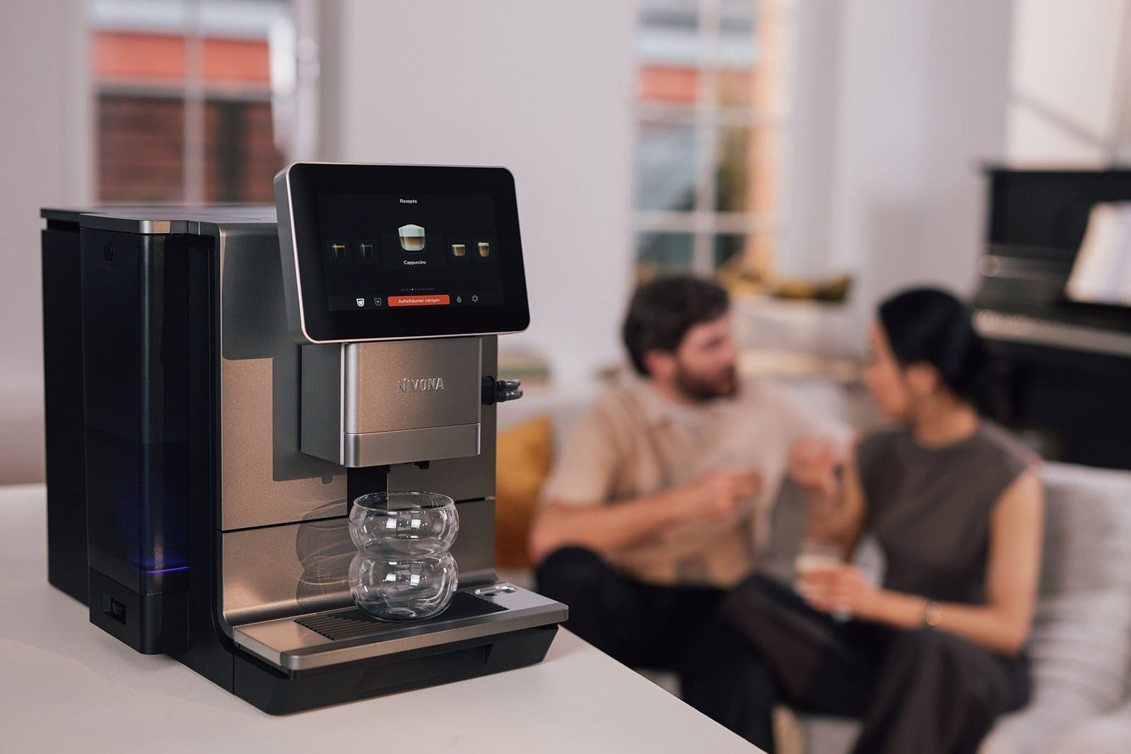 NIVONA Kaffeevollautomat der 9000er Serie auf einer Küchentheke, im Hintergrund ein Paar auf dem Sofa mit Kaffee